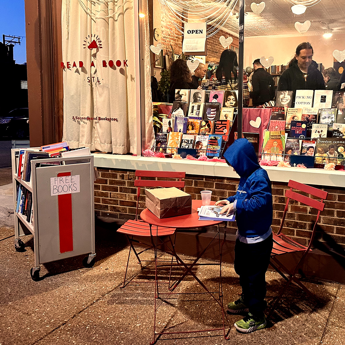 A child reads a book at a table on the sidewalk in front of Read A Book STL, a community-focused used bookstore at 3125 Meramec Street in Dutchtown, St. Louis, MO. Read A Book STL is a 2025 Dutchtown Façade Grant recipient.